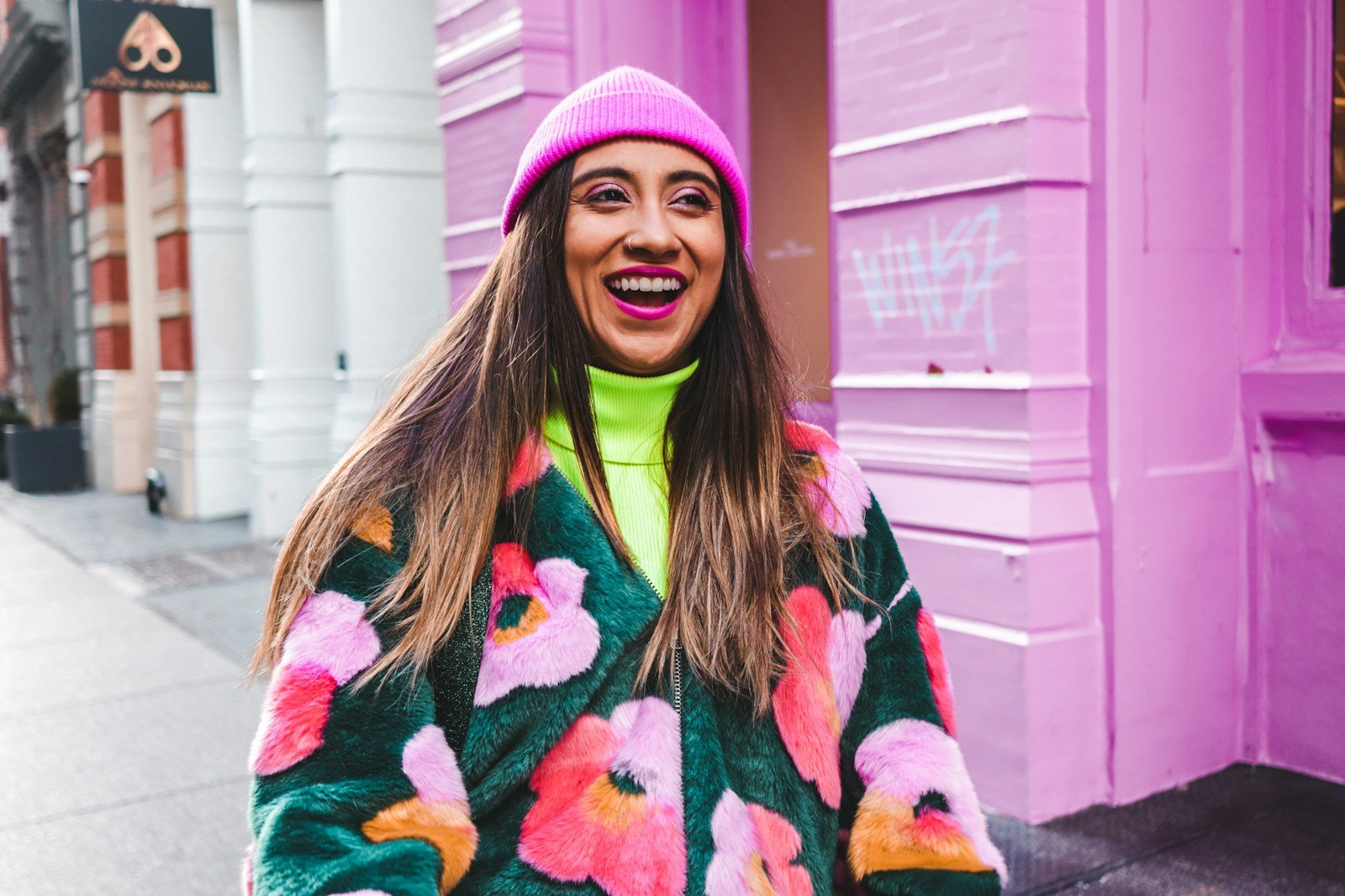 Laughing woman in a pink beanie and bright floral coat on a colourful street