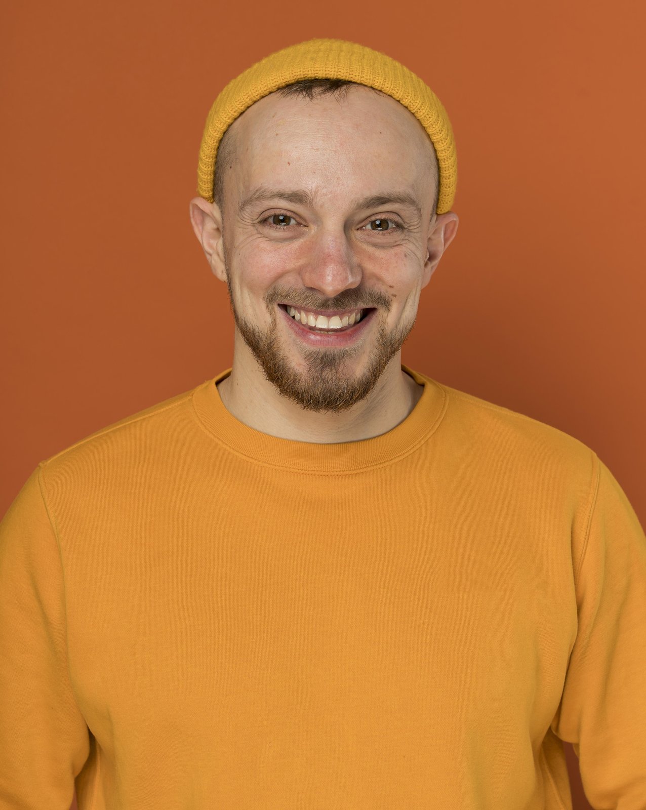 Bearded man in a yellow beanie grinning playfully at the camera